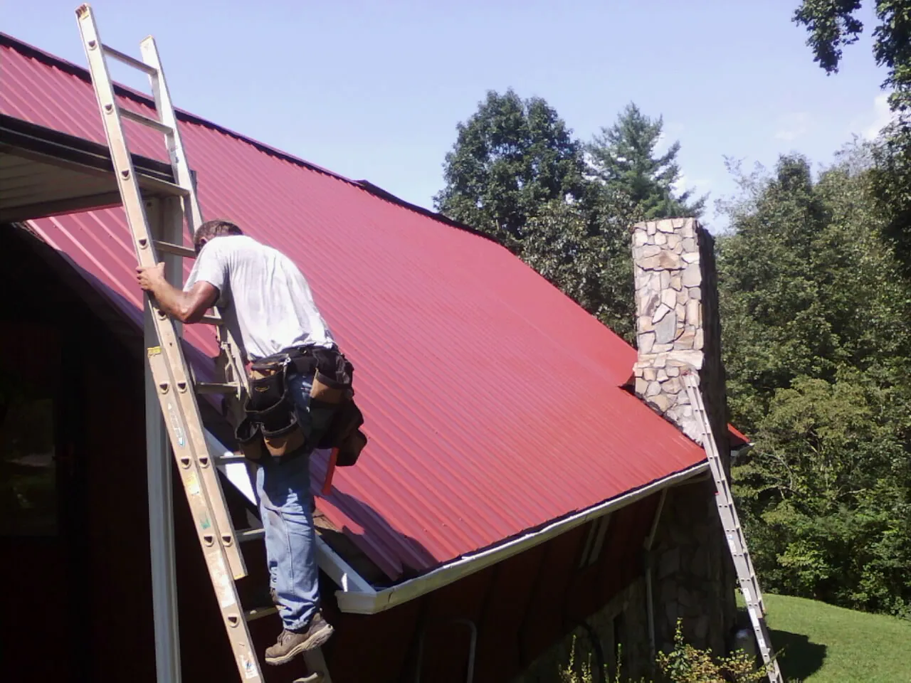 Roofer on a ladder beside a red metal roof and stone chimney — Soffit & Fascia Repair by Roofing Brothers in Essington